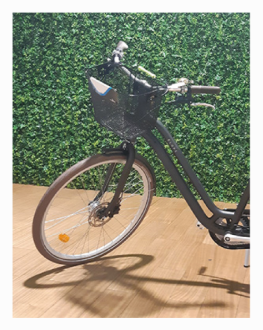 Fotografia da parte da frente de uma bicicleta cinzenta, com um cesto de rede preso ao guiador, sobre um chão de madeira e com uma parede de folhagem verde por trás. Crédito: «Imagem: © EduQA 2025».